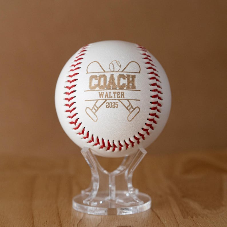 May include: A white baseball with red stitching, personalized with the word "COACH", the name "WALTER", and the year "2025". The baseball is displayed on a clear acrylic stand, showcasing the custom engraving.