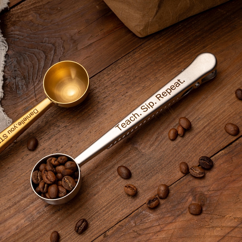 May include: A silver-toned metal coffee scoop with an integrated bag clip on the handle, engraved with the text "Teach. Sip. Repeat." The scoop is filled with roasted coffee beans. A second gold-toned scoop is partially visible in the upper left.