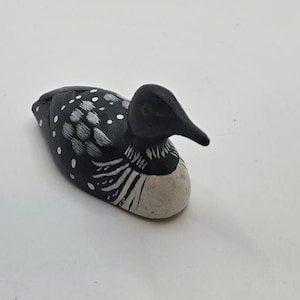 May include: A small hand-painted wooden loon figurine featuring a black head and back with white polka dots and striped markings on the neck and wings. The bird has a solid white chest and a pointed black beak, captured in a resting position.