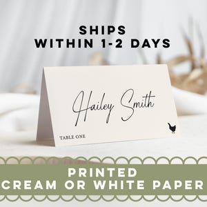 May include: A folded cream-colored paper place card featuring the black printed name Hailey Smith in a cursive script font, the text TABLE ONE in a sans-serif font, and a small black silhouette of a chicken on the bottom right corner.
