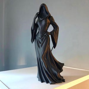 May include: A matte black figurine of a hooded figure wearing a long, flowing gown with wide, flared sleeves. The figure stands in a pose with one hand resting on the hip and the other near the hood, featuring a smooth, solid-colored surface.