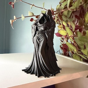 May include: A black figurine depicting two hooded figures in a close embrace. The figures are draped in long, flowing robes that reach the base, with skeletal hands visible as they hold each other. The sculpture has a smooth, matte finish.
