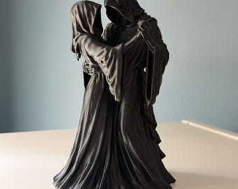 May include: A matte black figurine depicting two hooded figures in long, flowing robes engaged in a dance. The figures are draped in dark fabric with deep folds, and their faces are obscured by hoods, creating a unified, sculptural composition.