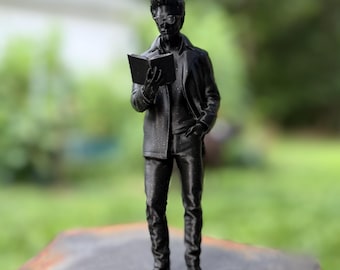May include: A black figurine of a person standing upright while holding an open book. The figure wears a collared jacket, a shirt, trousers, and glasses. The surface of the figurine has a subtle, fine-grained texture with a slight sheen.