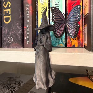 May include: A dark gray 3D printed figurine of a seated figure wearing a long robe and a pointed hat, featuring a textured surface. The figure is positioned in front of a large, iridescent purple and black butterfly ornament with white dots.
