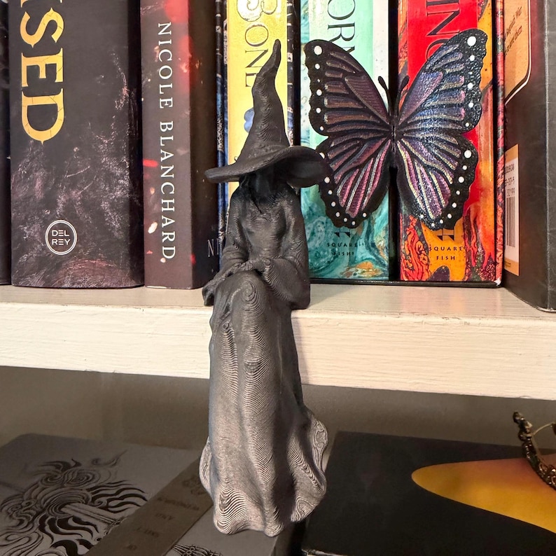 May include: A dark gray 3D printed figurine of a seated figure wearing a long robe and a pointed hat, featuring a textured surface. The figure is positioned in front of a large, iridescent purple and black butterfly ornament with white dots.