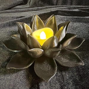 Puede incluir: Un portavelas negro con forma de flor de loto con una vela LED amarilla en el centro. El portavelas tiene dise&ntilde;os detallados de p&eacute;talos y se encuentra sobre una superficie oscura. Es un art&iacute;culo decorativo.