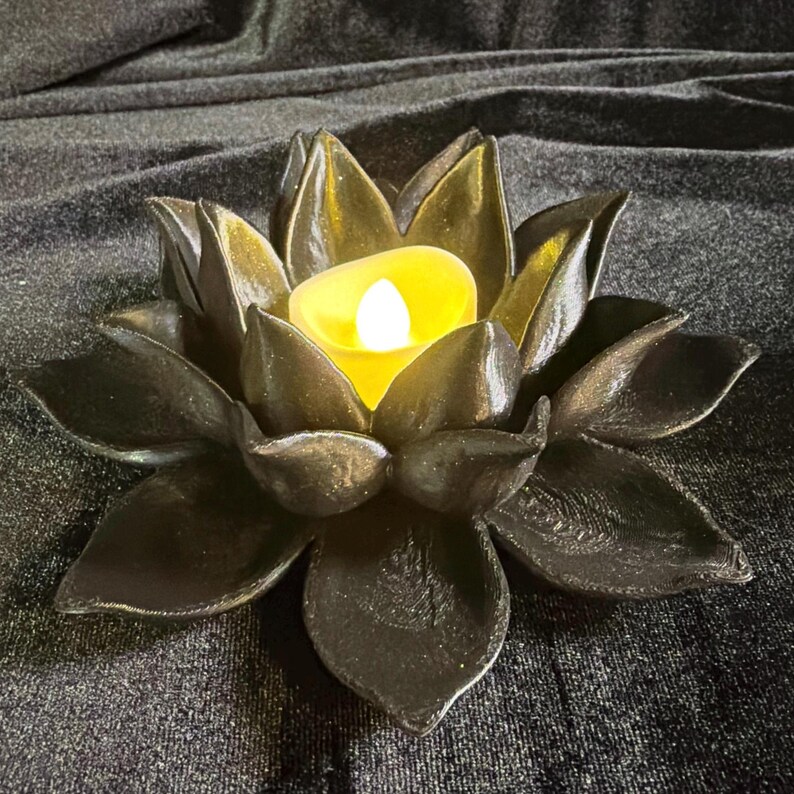 Puede incluir: Un portavelas negro con forma de flor de loto con una vela LED amarilla en el centro. El portavelas tiene dise&ntilde;os detallados de p&eacute;talos y se encuentra sobre una superficie oscura. Es un art&iacute;culo decorativo.