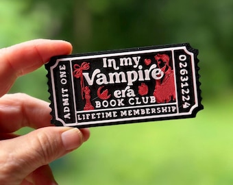 May include: A black rectangular ticket-shaped token with white and red raised lettering and graphics. It reads In my Vampire era BOOK CLUB, ADMIT ONE, and LIFETIME MEMBERSHIP, with a serial number 02631224, a chess piece, a heart, and an apple.