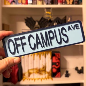 May include: A rectangular street sign with a light blue background and a dark blue border. The sign features raised dark blue lettering that reads OFF CAMPUS AVE in a sans-serif font. The sign has a clean, flat design with a smooth finish.