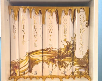 May include: A set of six white book spines arranged side-by-side, featuring gold foil lettering and a metallic gold drip effect covering the top and bottom edges. The titles include GILD, GLINT, GLEAM, GLOW, GOLD, and GOLDFINCH, each with small birdcage icons.