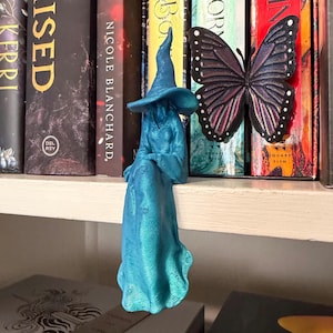 May include: A teal-colored 3D-printed figurine of a seated figure wearing a wide-brimmed pointed hat and a long, flowing robe. The figure is positioned on a shelf edge next to a decorative butterfly ornament with black, purple, and pink wings.
