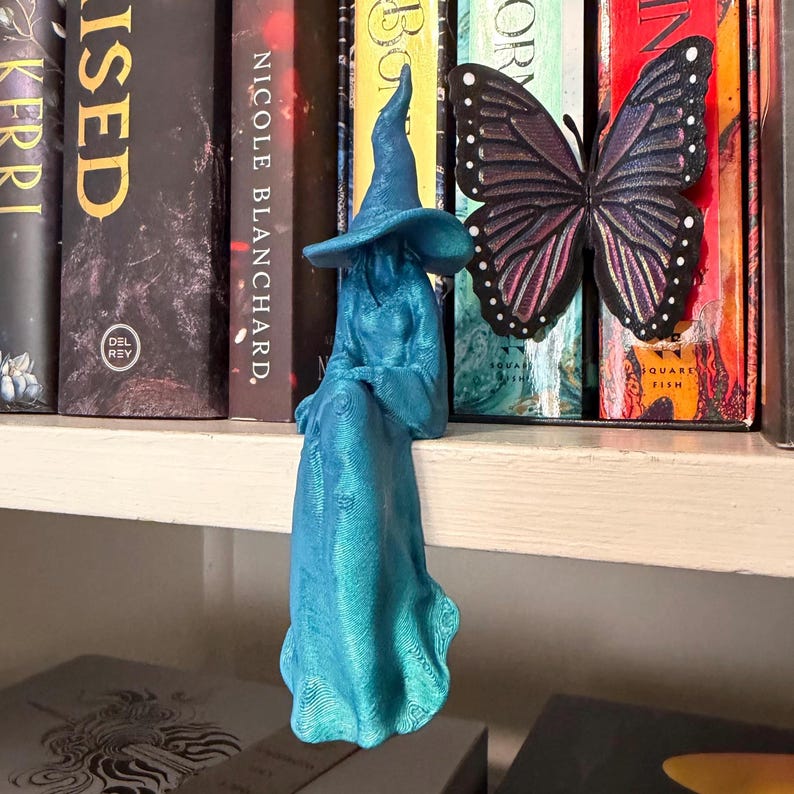 May include: A teal-colored 3D-printed figurine of a seated figure wearing a wide-brimmed pointed hat and a long, flowing robe. The figure is positioned on a shelf edge next to a decorative butterfly ornament with black, purple, and pink wings.