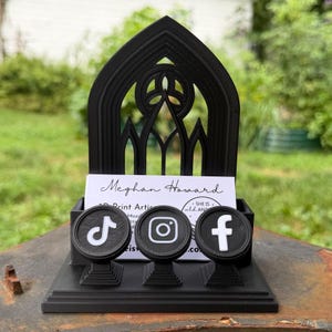 May include: A black 3D-printed display stand featuring a pointed gothic arch back panel. The base holds a white business card with the text Meghan Howard, 3D Print Artist, and She Is Wild And. Three circular icons for TikTok, Instagram, and Facebook are attached.