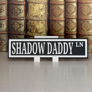 May include: A black and white street sign that reads "SHADOW DADDY LN". The sign is rectangular and has white lettering on a black background. It is supported by two white stands. The background features a stack of antique books.