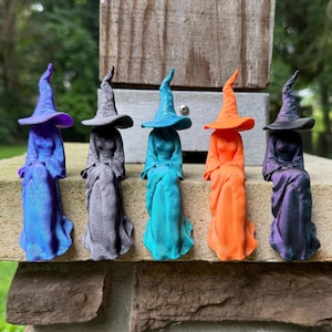 May include: Five small witch figurines in a row, each wearing a pointed hat and a long dress. The figures are colored in iridescent blue, matte gray, bright teal, vibrant orange, and a shimmering purple-black gradient, all seated upright.