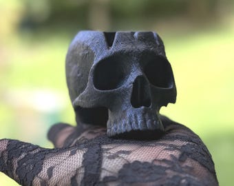 May include: A matte black skull-shaped container with a hollow top. The skull features deep, dark eye sockets, a defined nasal cavity, and visible teeth detailing. The surface has a textured, slightly rough finish, giving it a dark appearance.