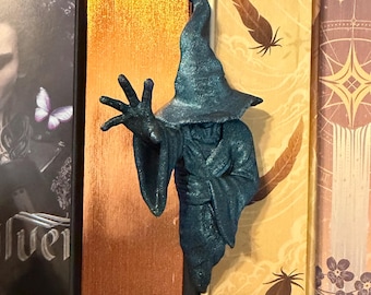 May include: A dark teal, textured figurine of a robed figure wearing a pointed wizard hat. The figure is depicted with one arm extended forward, palm open, and fingers spread wide. The surface has a subtle, shimmering, granular finish.