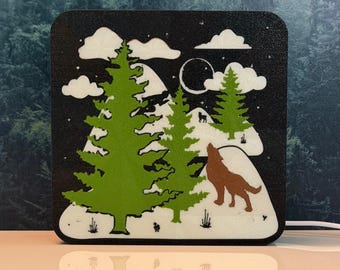 May include: A square decorative light panel featuring a black background with a white moon, clouds, and snow-covered mountains. Two bright green pine trees stand in the foreground, while a brown wolf silhouette is positioned on a snowy hill.