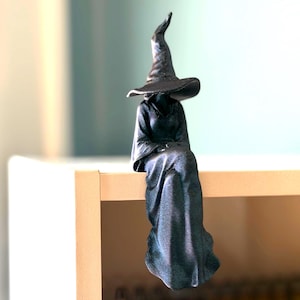 Puede incluir: Una figura negra de un personaje sentado que lleva una túnica larga y fluida y un sombrero alto y puntiagudo. La figura tiene una superficie texturizada y está colocada para sentarse en el borde de una superficie plana, con la túnica colgando.