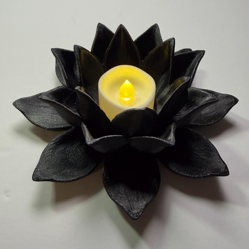 Puede incluir: Un portavelas de flor de loto negro con una vela que funciona con pilas. La vela tiene una llama amarilla parpadeante. Los p&eacute;talos de la flor son texturizados y rodean la vela, creando un aspecto decorativo y elegante.