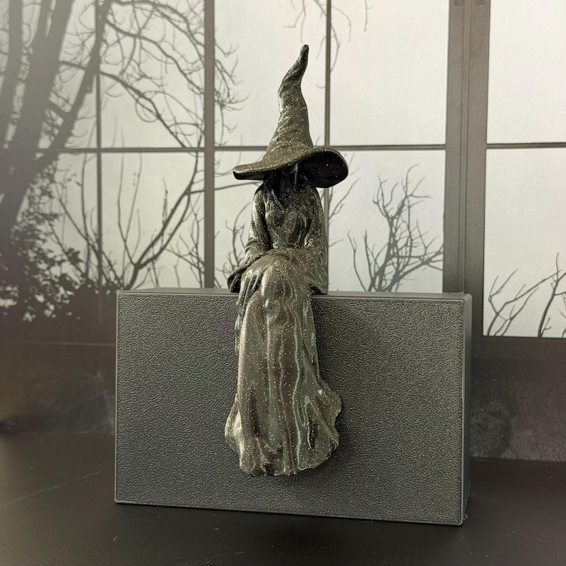 May include: A dark, glittery figurine of a seated figure wearing a tall, wide-brimmed pointed hat and a long, flowing robe. The figure is positioned on the edge of a rectangular, textured dark gray block, creating a shelf-sitting display.