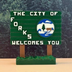 以下が含まれることがあります： THE CITY OF FORKS WELCOMES YOUと書かれた白い浮き彫り文字がある緑色の長方形の看板。看板には木とトラックが描かれた青、白、茶色の円形エンブレムがあり、緑色の台座の上に2本の茶色の支柱で支えられています。
