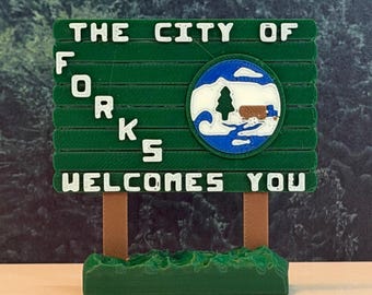 May include: A green rectangular sign with white raised lettering that reads THE CITY OF FORKS WELCOMES YOU. The sign features a circular emblem with a blue, white, and brown graphic of a tree and a truck, supported by two brown posts on a green base.