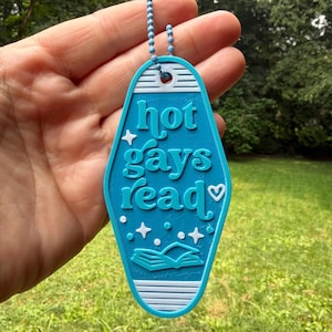 May include: A teal motel-style keyring with white raised text that reads hot gays read, featuring a small white heart, four-pointed stars, and an open book graphic. The keyring has a white border and a blue ball chain attached at the top.
