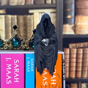 May include: A black figurine of a hooded skeleton figure draped in a long robe, holding a small white cup with skeletal hands. The figure is positioned on top of a book spine, next to a gold crown featuring large green gemstones.