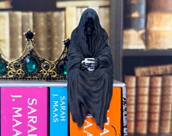 May include: A black figurine of a hooded skeleton figure draped in a long robe, holding a small white cup with skeletal hands. The figure is positioned on top of a book spine, next to a gold crown featuring large green gemstones.