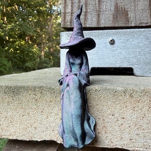 May include: A seated witch figurine with a tall, pointed hat and a long, flowing robe. The surface of the sculpture features a distinct, layered texture with a color-shifting finish that transitions between shades of deep purple, blue, and teal.