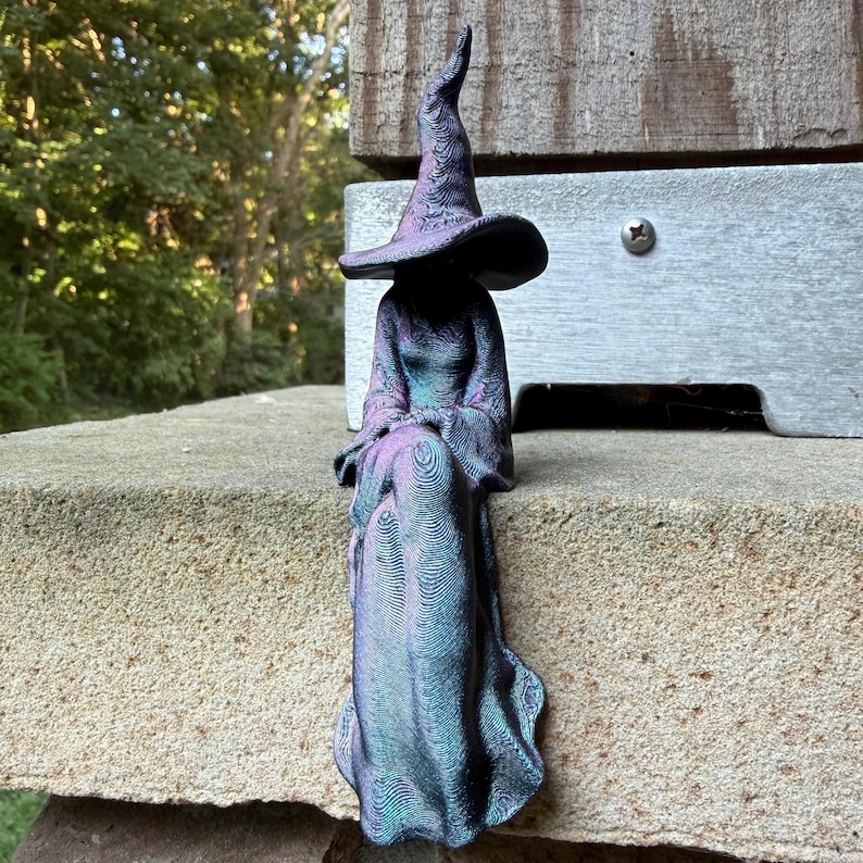 May include: A seated witch figurine with a tall, pointed hat and a long, flowing robe. The surface of the sculpture features a distinct, layered texture with a color-shifting finish that transitions between shades of deep purple, blue, and teal.