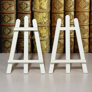 May include: Two small, white wooden easels. Each easel has a three-legged design with a horizontal support bar. The background features a collection of antique books with aged, textured covers.