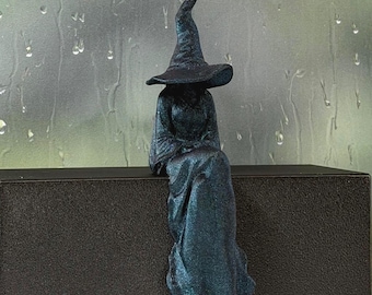May include: A dark blue, glittery figurine of a seated figure wearing a tall, pointed hat and a long, flowing robe. The figure is positioned with its legs hanging down, featuring a textured surface that catches the light with subtle sparkles.