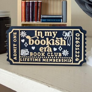 May include: A navy blue and cream-coloured decorative ticket with the text "In my bookish era BOOK CLUB LIFETIME MEMBERSHIP." The ticket has floral and heart designs, and the words "ADMIT ONE" are printed on the left side.