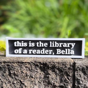 May include: A rectangular sign with a white border and a black background featuring the white text "this is the library of a reader, Bella" in a bold, sans-serif font. The sign is oriented horizontally with a clean, high-contrast design.