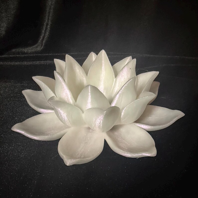 Puede incluir: Una flor de loto blanca con m&uacute;ltiples p&eacute;talos, que exhibe un sutil brillo iridiscente. La flor est&aacute; centrada y en plena floraci&oacute;n, con un suave brillo nacarado. Los p&eacute;talos est&aacute;n en capas, creando una apariencia detallada y texturizada.
