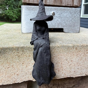 May include: A black figurine of a seated witch wearing a tall, pointed hat and a long, flowing gown. The surface of the figure has a textured, speckled appearance with visible fine lines throughout the material, depicting a draped garment.