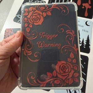 Puede incluir: Una funda transparente para teléfono con fondo negro y diseño floral rojo. El diseño incluye rosas y la frase "Trigger Warning". La funda está sujeta en la mano de una persona.