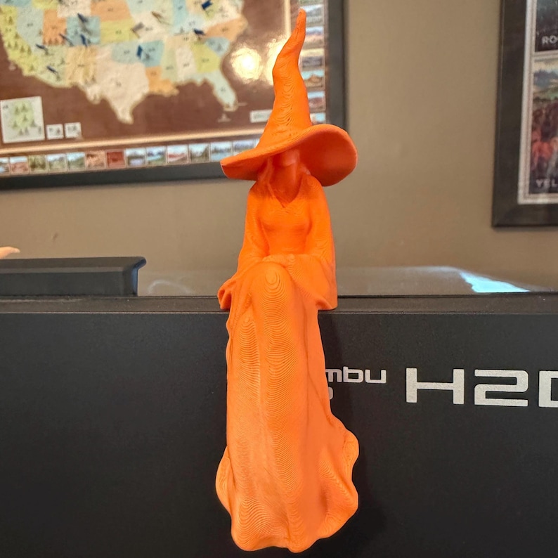 May include: A bright orange 3D printed figurine of a witch wearing a tall, pointed hat and a long, flowing gown. The figure is designed to sit on an edge, with the skirt draping downwards, showcasing visible horizontal layer lines from the printing process.