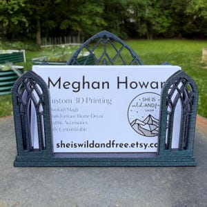 May include: A dark teal, glittery 3D-printed business card holder featuring a gothic arch design. It holds a white business card with the text Meghan Howard, Custom 3D Printing, Bookish Magic, Dark Fantasy Home Decor, Gothic Accessories, Fully Customizable, and sheiswildandfree.etsy.com.