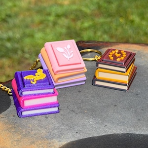 May include: Three miniature book keychains with gold-tone metal rings. One stack features a purple book with a yellow dragon emblem, another has a pink book with a white floral design, and the third is a dark red book with a yellow heart pattern.