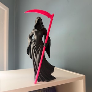 May include: A matte black figurine of a cloaked figure with a hooded head, wearing a long flowing robe with wide sleeves. The figure holds a tall, bright neon pink scythe that extends from the base to above the head of the hooded character.