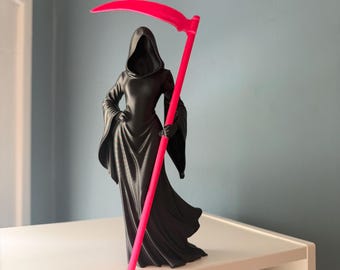May include: A matte black figurine of a cloaked figure with a hooded head, wearing a long flowing robe with wide sleeves. The figure holds a tall, bright neon pink scythe that extends from the base to above the head of the hooded character.
