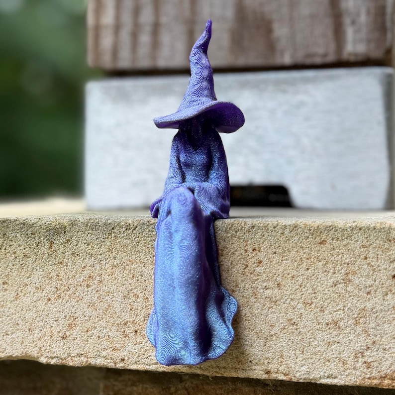 May include: A purple 3D printed figurine of a seated figure wearing a long, flowing robe and a tall, pointed hat. The surface of the figure displays a visible, fine-lined texture throughout the garment and hat, with a subtle shimmering finish.