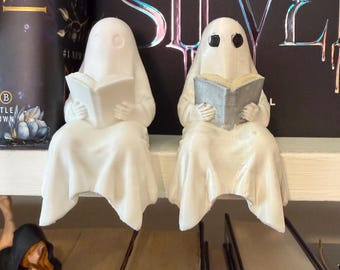 May include: Two white ghost figurines, each reading a book, sit on a shelf. The ghosts are draped in white fabric, with simple eye cutouts. One book is white, the other is gray. The background includes book spines.