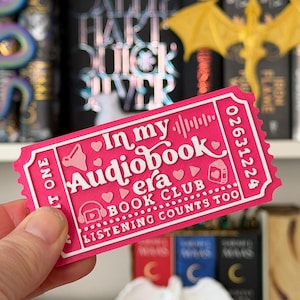 May include: A pink ticket-shaped sticker with the text "In my Audiobook era BOOK CLUB LISTENING COUNTS TOO". The sticker has a white border and features icons of a speaker, headphones, and a book.