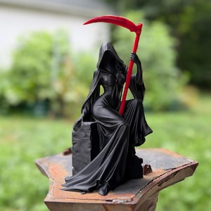 May include: A black figurine of a hooded figure in a long flowing robe sitting on a stone-like block. The figure holds a tall, bright red scythe that extends upward. The sculpture features smooth, dark surfaces with sharp, vibrant red accents.