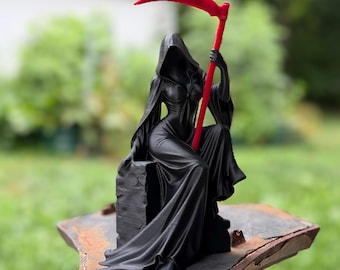 May include: A black figurine of a hooded figure in a long flowing robe sitting on a stone-like block. The figure holds a tall, bright red scythe that extends upward. The sculpture features smooth, dark surfaces with sharp, vibrant red accents.